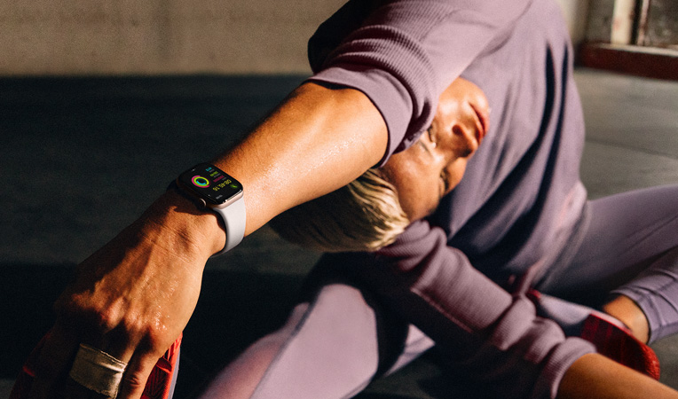 一位女士戴着 Apple Watch Series 10 坐在地板上，手臂向腿部进行拉伸，屏幕上显示体能训练 app 中的整理放松、健身记录和健身圆环。